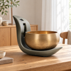 An automatic brass singing bowl resting in a curved matte gray holder on a wooden table, with a softly blurred indoor room in the background.